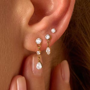 Könnte beinhalten: Nahaufnahme eines Ohrs, das mit mehreren goldenen Ohrringen geschmückt ist. Die Ohrringe verfügen über funkelnde, klare Edelsteine in verschiedenen Größen und Formen, darunter runde und tropfenförmige Designs. Der Schmuck ist vor dem Hintergrund der Haut und der Haare einer Person platziert.