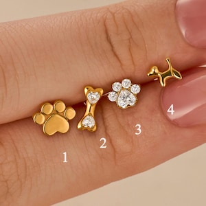May include: Four gold-coloured stud earrings are displayed. The earrings include a paw print, a dog bone with clear stones, a flower-shaped design with clear stones, and a small dog silhouette. The earrings are presented against a light background.