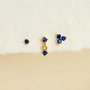 Op de afbeelding: Een set van drie goudkleurige oorbellen met kleine, ronde, donkerblauwe stenen. De set bevat een knopje met één steen, een hangende oorbel met twee stenen verbonden door een ringetje, en een knopje met drie stenen in een cluster geplaatst.