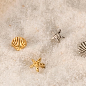 May include: A selection of beach-themed jewellery. The image shows two gold and two silver pieces, including two seashell-shaped studs and two starfish-shaped studs. The jewellery is displayed on white sand.