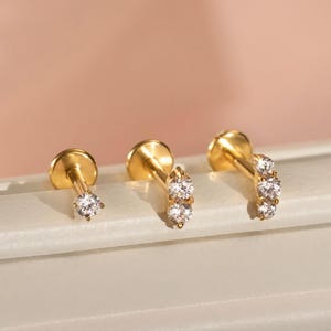 May include: Three gold-tone stud earrings featuring clear, round-cut stones. The first earring has a single stone, the second has two stones in a vertical row, and the third has three stones arranged in a curved, vertical line on a post back.