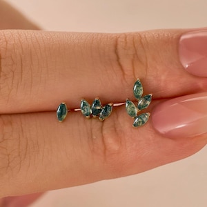 May include: A delicate gold earring featuring a leaf design with multiple green gemstones. The gemstones are oval-shaped and arranged to mimic leaves on a branch. The earring is held up by a person's fingers.