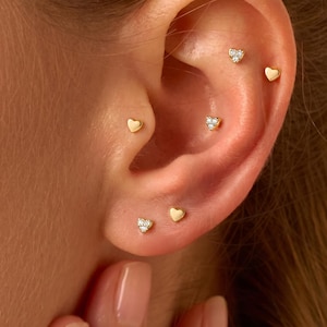 May include: A set of five small gold-toned earrings worn in a single ear. The collection includes three heart-shaped studs with tiny embedded clear stones and two plain, smooth gold-toned heart-shaped studs arranged along the ear cartilage and lobe.