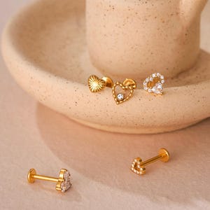 May include: Several gold-coloured earrings, featuring heart and sunburst designs, are arranged on a beige ceramic dish. Some earrings are embellished with small, clear gemstones. The earrings are presented on a pink cloth.