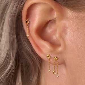 May include: Gold-tone earrings featuring delicate chains with small moon and star charms. One earring is placed in the upper cartilage, while two earrings are positioned in the earlobe. Each charm is accented with a tiny, clear, sparkling stone.