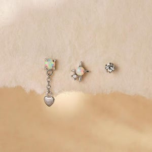 Peut inclure: Trois boucles d'oreilles à tige argentées. La première présente une pierre opale avec une breloque cœur pendante. La deuxième est un sertissage en forme d'étoile avec une opale centrale et de petites pierres claires. La troisième est un petit clou rond en pierre claire. Le texte indique Gleamy Day.