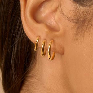 May include: Three gold hoop earrings of different sizes are shown in the ear. The earrings have a shiny, polished finish, reflecting light. The hoops are a simple, classic design, suitable for everyday wear.