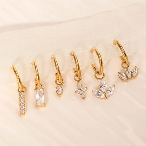 Peut inclure: Une collection de six boucles d'oreilles créoles dorées avec divers breloques pendantes. Les breloques comprennent une barre verticale de petites pierres transparentes, une pierre rectangulaire transparente, une pierre en forme de goutte, une fleur à quatre pétales et un design à trois pétales.