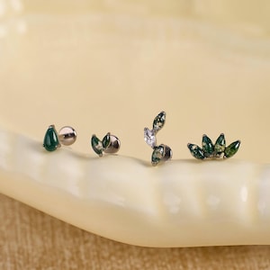 May include: Four individual stud earrings featuring dark green teardrop and marquise-cut stones set in silver-toned metal. The designs include a single teardrop, a double leaf, a curved vine with a clear stone, and a five-stone fan-shaped cluster.
