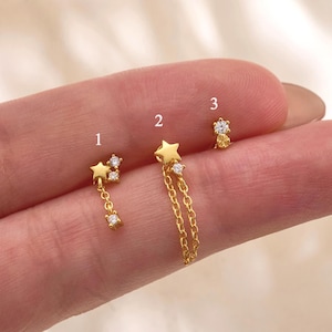 May include: Three gold-tone stud earrings labeled 1, 2, and 3. Earring 1 features a star and a dangling chain with a clear stone. Earring 2 has a star and a double chain with a clear stone. Earring 3 is a small stud with a clear stone. Text: Gleamy Day.