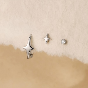 May include: A set of three silver-toned earrings consisting of one star-shaped hoop, one small four-pointed star stud, and one round stud with a clear stone. The text Gleamy Day is printed at the bottom of the image.