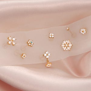 May include: A selection of gold-coloured stud earrings featuring diverse floral and geometric patterns. The earrings are embellished with tiny, sparkling crystals. The earrings are presented on a sheer, white fabric over a pink satin backdrop.