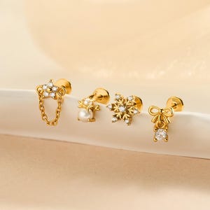 May include: Four gold stud earrings with different designs. One has a chain and gem design, another a pearl, a snowflake, and a bow with a gem. The earrings are displayed on a white, curved surface against a soft, neutral background.