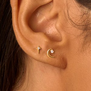 May include: A close-up of an ear with two gold earrings. One is a starburst design, and the other is a crescent moon shape. Both earrings feature a small, clear gemstone. The earrings are set against the backdrop of the ear and surrounding skin.
