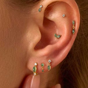 May include: An ear with multiple gold earrings, each featuring emerald green gemstones and clear crystals. The earrings include studs, drop styles, and small hoops. The image also shows the text "Gleamy Day" at the bottom.