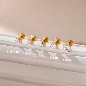 Peut inclure: Cinq boucles d'oreilles à tige dorées avec des pierres rondes transparentes, disposées en rangée de la plus petite à la plus grande. Les tailles sont indiquées sous chaque clou comme 1,5 mm, 2 mm, 2,5 mm, 3 mm et 4 mm pour le diamètre.