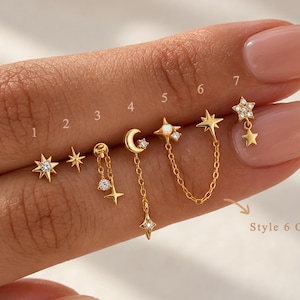 May include: A collection of seven gold-toned stud earrings labeled one through seven, featuring celestial motifs including stars, a crescent moon, and small clear stones. Some designs incorporate delicate gold chains and an opal-like accent stone.