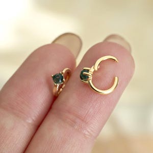 May include: Two gold-coloured hoop earrings, each featuring a small, round, dark teal gemstone. One earring is closed, while the other is open, showcasing the clasp mechanism. The earrings are displayed against a soft, neutral background.
