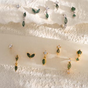 May include: A collection of earrings with green and clear gemstones. The designs include studs, drop earrings, and hoops, with leaf and floral motifs. The earrings are displayed on a textured, off-white surface.