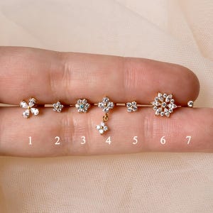 May include: A collection of seven small, flower-shaped earrings with clear gemstones. The earrings are arranged in a row, showcasing different designs and sizes. The earrings are set on a light-coloured background.
