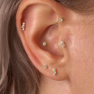 May include: Close-up of an ear with multiple gold earrings. The earrings feature small, sparkling gemstones in various designs, including single studs and clusters of two or three stones. The ear is set against a background of light-coloured hair.