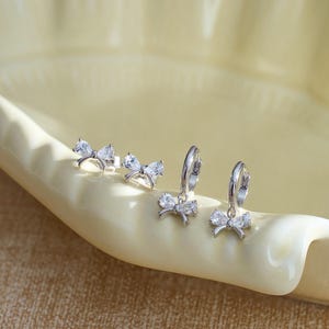Peut inclure: Plusieurs boucles d'oreilles argentées avec des motifs de nœuds, comprenant des modèles à clous et à créoles. Les nœuds sont ornés de pierres précieuses claires et étincelantes. Les boucles d'oreilles sont présentées sur un plat en forme de feuille jaune clair.