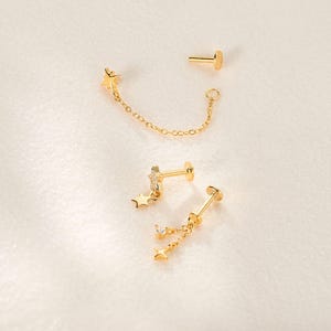 May include: Three gold-tone stud earrings with flat back posts. One features a star connected by a delicate chain to a ring. Another is a star-shaped stud with a small dangling star. The third is a stud with two dangling stars and a clear stone.