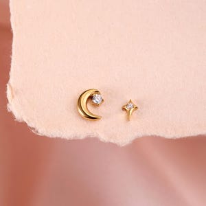 May include: A pair of gold-toned stud earrings consisting of a crescent moon shape with a single clear round stone and a four-pointed star shape with a single clear round stone at the center of each piece.