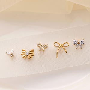 May include: An assortment of gold-toned stud earrings in various bow and butterfly designs. Some are adorned with clear crystals, while others have a solid gold appearance. The earrings are arranged on a cream-colored fabric.