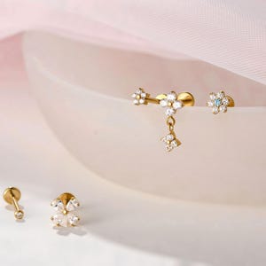 May include: Gold-coloured earrings with floral designs, set with small, clear gemstones. The earrings are displayed on a white, curved surface against a soft pink background. Styles include studs and dangle earrings.