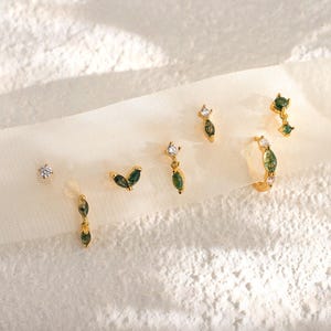 Op de afbeelding: Een collectie gouden oorbellen met verschillende ontwerpen, waaronder studs, bungelende stijlen en een oorring. De oorbellen zijn voorzien van groene en heldere edelstenen. De afbeelding is geplaatst tegen een zachte, witte achtergrond. De tekst "Gleamy Day" is zichtbaar.