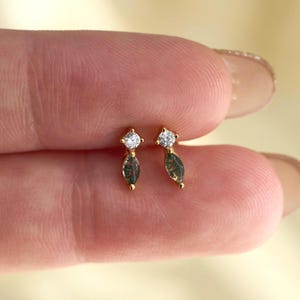 Könnte beinhalten: Ein Paar goldene Ohrstecker. Jeder Ohrring hat oben einen klaren, rund geschliffenen Stein und unten einen dunkelgrünen, marquiseförmigen Stein. Die Ohrringe sind klein und zart, mit einem minimalistischen Design.