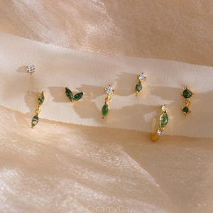 May include: Several gold earrings with different designs, including studs, leaf shapes, and green teardrop stones. Some earrings have clear, sparkling details. The earrings are displayed on a soft, cream-coloured fabric.