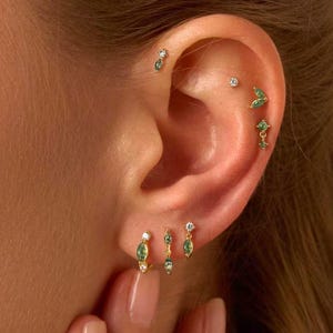 May include: An ear displaying multiple gold earrings with small green gemstones and clear crystals. The ear is pierced in several places, showcasing a variety of delicate jewelry designs. The text "Gleamy Day" is visible at the bottom of the image.