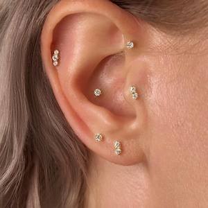 Könnte beinhalten: Nahaufnahme eines Ohrs, das mit mehreren goldenen Ohrringen geschmückt ist. Die Ohrringe weisen kleine, funkelnde Edelsteine in verschiedenen Designs auf, darunter einzelne Ohrstecker und Cluster aus zwei oder drei Steinen.