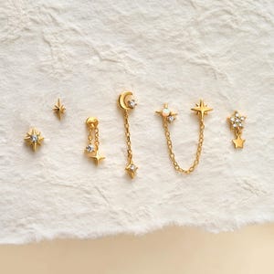 May include: A set of gold-toned earrings featuring celestial motifs including a starburst, a crescent moon with a dangling star, and a double-stud chain connector with a small opal-like stone and star accents, all adorned with clear crystals.