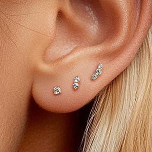 May include: Close-up of an ear with three small, sparkling stud earrings. The earrings are set with clear gemstones, and are arranged in a graduated style. The ear is set against a background of blonde hair.