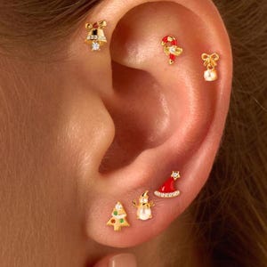 May include: An ear featuring multiple gold earrings with festive Christmas designs. The earrings include a Christmas tree, a snowman, a Santa hat, a bell, a candy cane, and a bow with a pearl. Each earring is decorated with tiny gemstones.