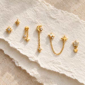 Peut inclure: Une collection de boucles d'oreilles dorées présentant des motifs célestes, notamment des étoiles, un croissant de lune et de petites pierres claires. Une boucle possède une chaîne reliant deux étoiles, d'autres ont des pampilles. Texte Gleamy Day.