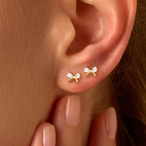 Peut inclure: Gros plan sur une oreille ornée de deux petites boucles d'oreilles en forme de nœud papillon dorées. Chaque boucle d'oreille présente une pierre transparente et étincelante au centre du nœud. Les boucles d'oreilles sont posées sur la peau, soulignant leur design délicat.