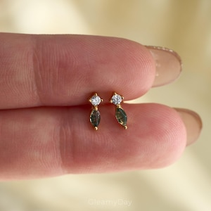 May include: A pair of gold stud earrings. Each earring has a clear, round gemstone at the top and a dark green, marquise-shaped stone below. The earrings are set against a soft, neutral background.