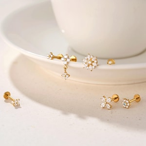May include: A selection of gold-coloured earrings with floral and geometric patterns. Several earrings are adorned with clear, sparkling gemstones. The earrings are presented on a white surface, highlighting their intricate details and elegant style.