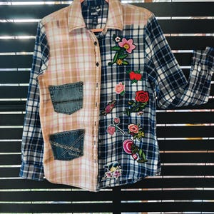 May include: A long-sleeve button-down shirt featuring a split design with one side in a bleached tan plaid and the other in a navy and white plaid. The shirt includes two denim pockets on the tan side and multiple colorful floral patches on the navy side.