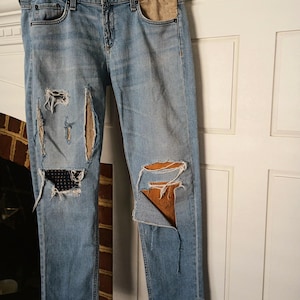 May include: Light blue denim jeans with distressed details, including rips and patches. The jeans have a classic five-pocket design and are hanging on a white hanger. The patches are a tan colour and the jeans are cuffed at the ankles.