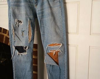 May include: Light blue denim jeans with distressed details, including rips and patches. The jeans have a classic five-pocket design and are hanging on a white hanger. The patches are a tan color and the jeans are cuffed at the ankles.