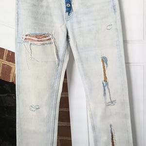 May include: Light wash denim jeans with distressed details, including rips and patches. The jeans feature a button fly and decorative pocket accents. The jeans are hanging on a clothesline.