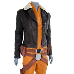 Może przedstawiać: Brązowa skórzana kurtka z kremowym kołnierzem z kożucha jest noszona na pomarańczowym kombinezonie. Strój zawiera brązowy pasek z kieszeniami i srebrną klamrą. Kostium jest prezentowany na manekinie.