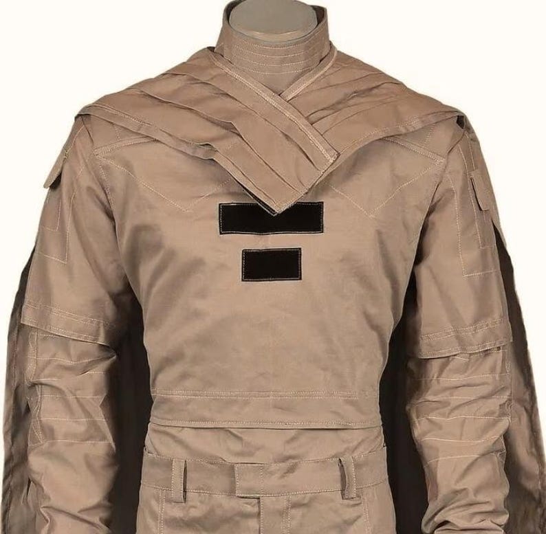 May include: Tan-colored military-style uniform with a high collar and a layered shoulder design. The uniform features two black rectangular patches on the chest and multiple pockets. The fabric appears to be a durable, possibly water-resistant material.