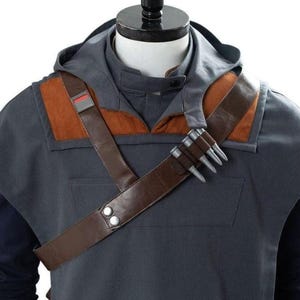 May include: A gray hooded jacket with brown leather shoulder straps and accents. The jacket features a brown leather strap with silver studs and a small red and gray patch. The jacket is part of a costume.