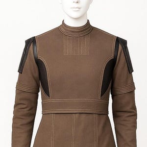 May include: A brown jacket with black leather panels on the shoulders and sides. It features a high collar, vertical stitching, and a fitted waist. The sleeves have a double-layered design. The jacket is displayed on a mannequin.